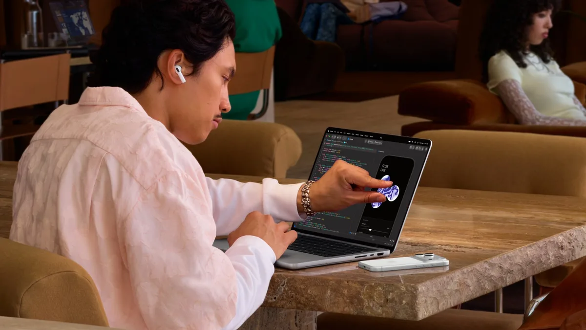 A person at a café table touching the screen of a MacBook Pro with PROLED XDR Display